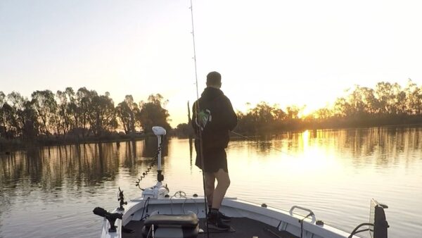A person fishing from the front of the boat with the sun setting over a lake surrounded by trees in the background