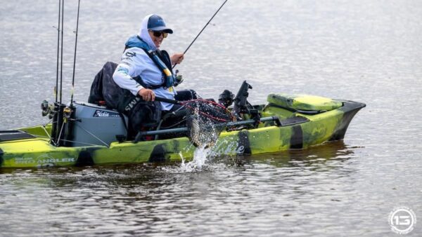A woman fishing in a green kayak with a fishing rod in one hand, using their other hand to net a fish