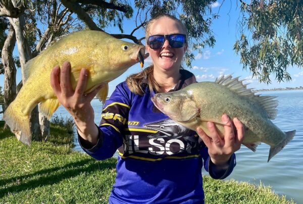 WIRFAB leader Kelsi Gull holding two golden perch by a grassy lake edge wearing a blue shirt